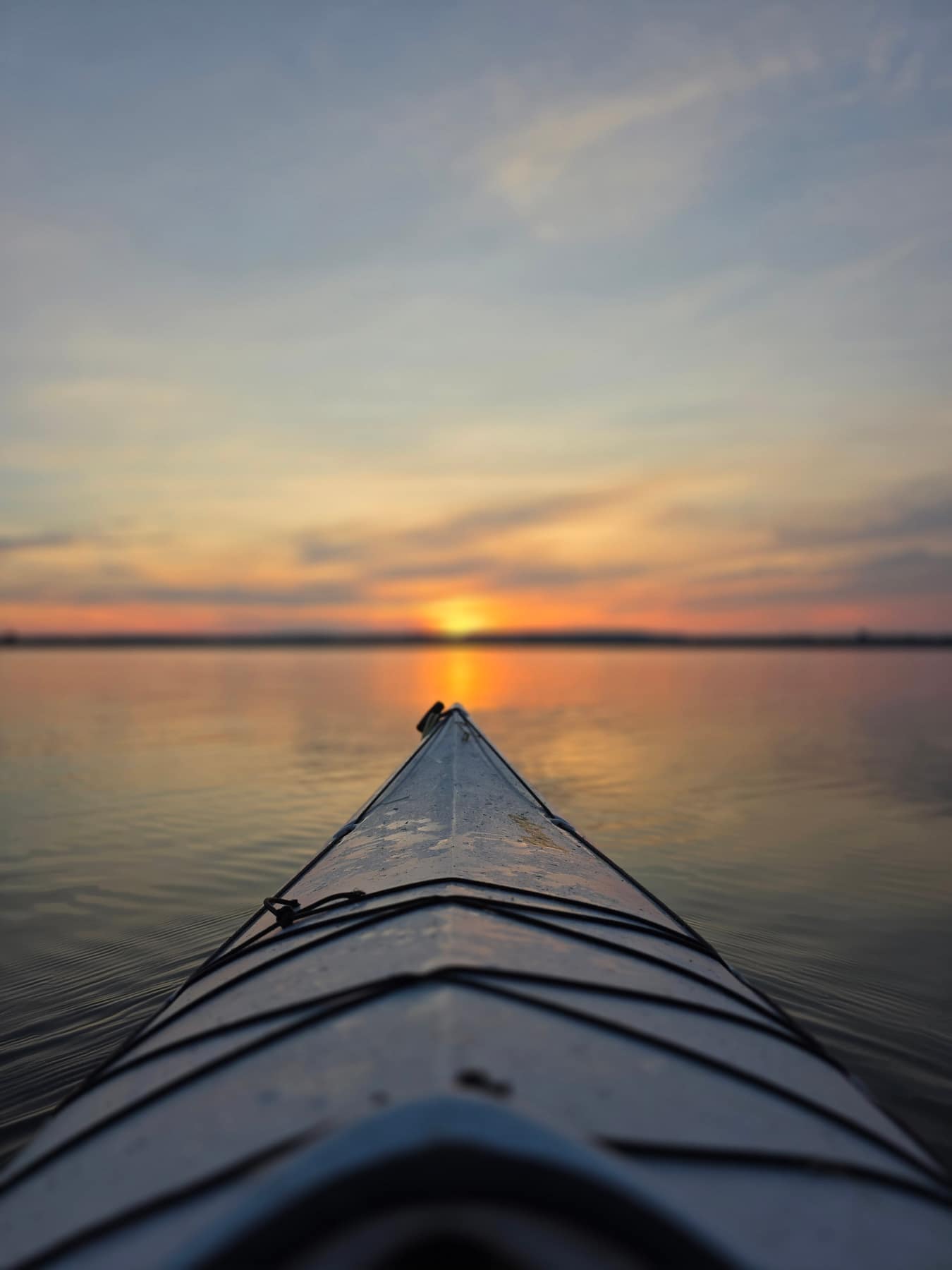 The bow of a kayak pointed straight at the sunset across flat water.