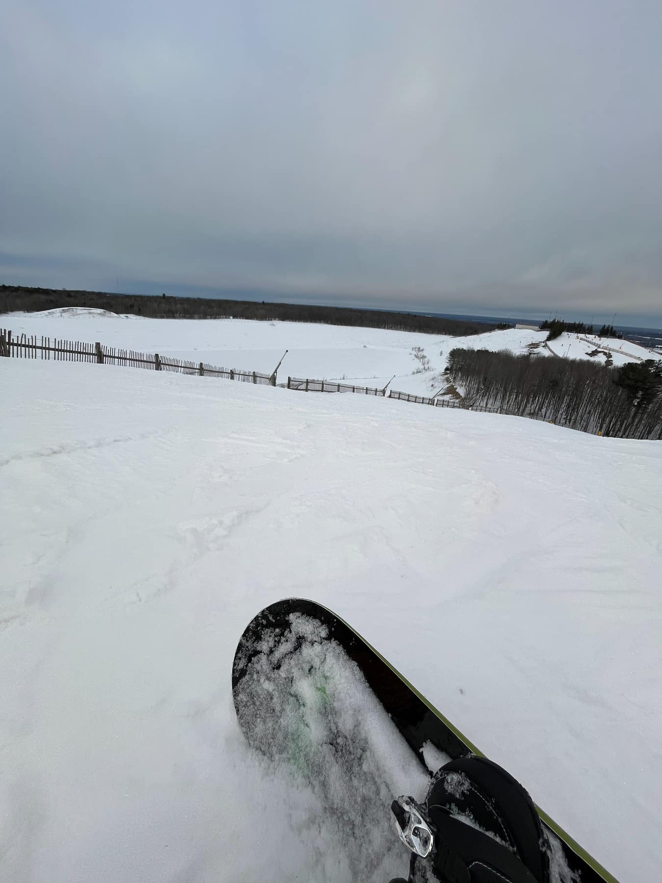 The nose of a snowboard at the top of a run, flat grey cloud over the hill.